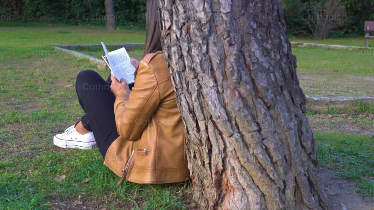 Mujer joven lee junto a un árbol, toma 2 - Banco de vídeos católicos - Fotografía por Cathopic
