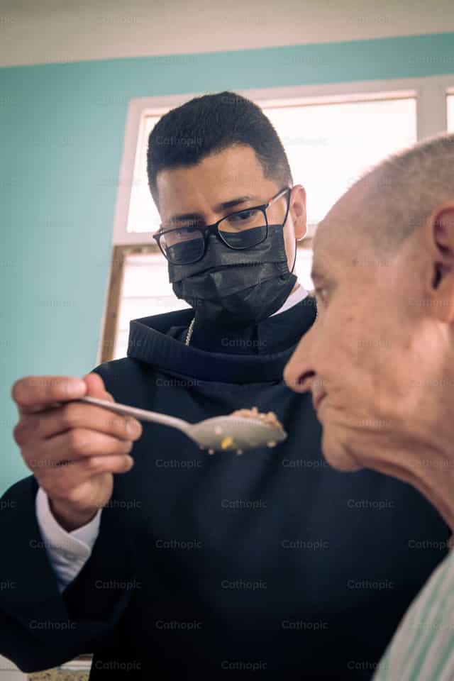Sacerdote con la mascherina serve un anziano - Immagini stock cattoliche