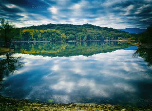 See mit spiegelndem, wolkenverhangenem Himmel und grünem Wald - Katholische Stockfotos