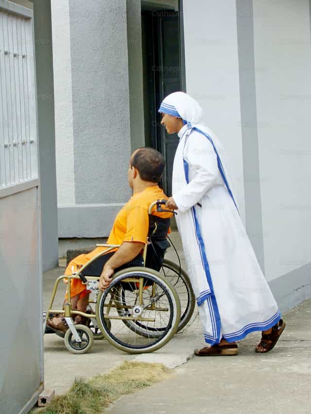 Una monja sonriente ayuda a una persona en silla de ruedas - Banco de imágenes católicas