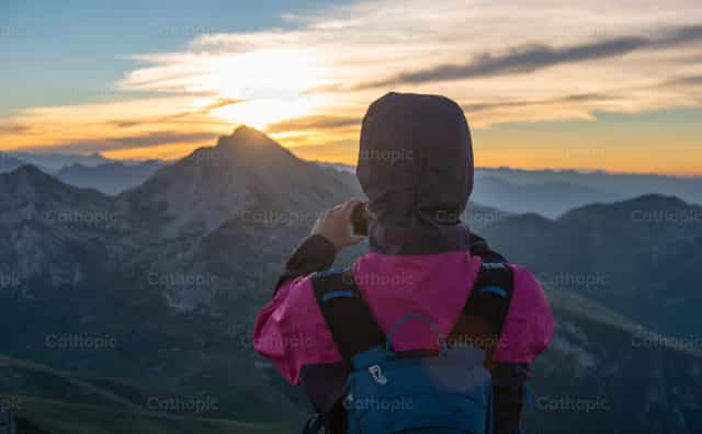 A hiker contemplates and photographs the sunrise amidst the mountains, admiring Creation.. - Catholic stock images - Photo by Pierluigi Palazzi