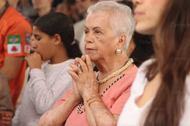 Una mujer mayor con las manos entrelazadas en oración, mirando fijamente al frente - Banco de imágenes católicas - Fotografía por Thácio Leite